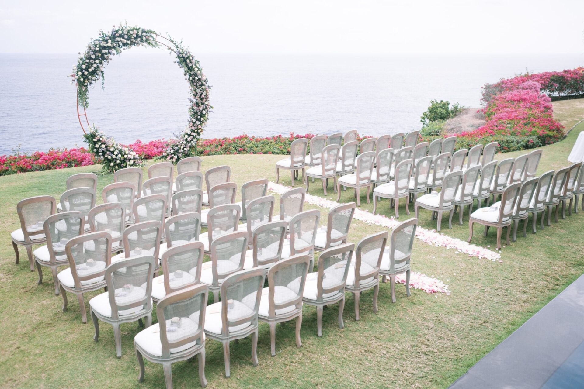 A wide shot of an arch for a wedding decorated with flowers and white chairs surrounded by beautiful pink flowers on the beach
